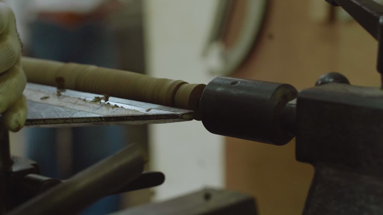 A gloved hand uses a chisel to shape a spinning wooden piece on a lathe, with fine wood shavings flying, capturing a detailed moment of skilled woodturning.