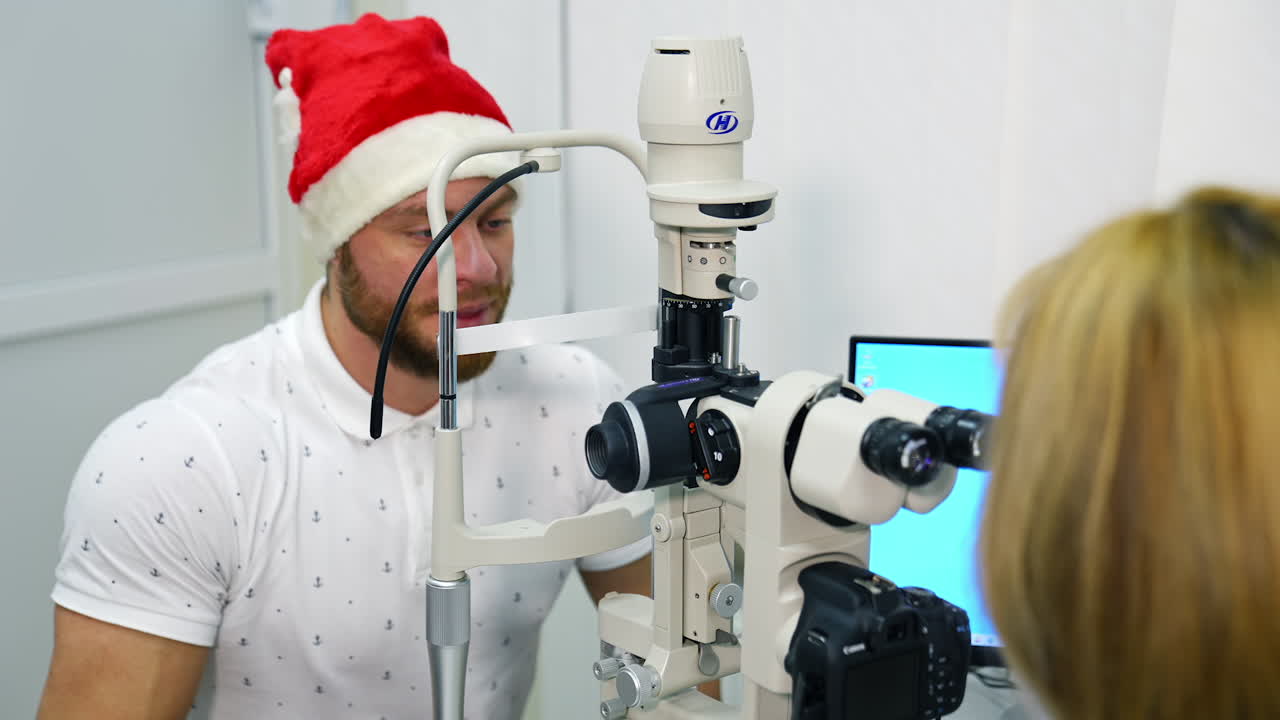 Doctor examining eye with medical equipment. Ophthalmologist check eyesight of young man in christmas cap