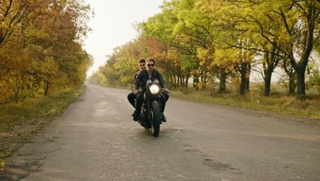 Handsome-man-in-sunglasses-riding-a-motorcycle-with-his-girlfriend-behind,-traveling-together-on-the-asphalt-road-in-forest-in