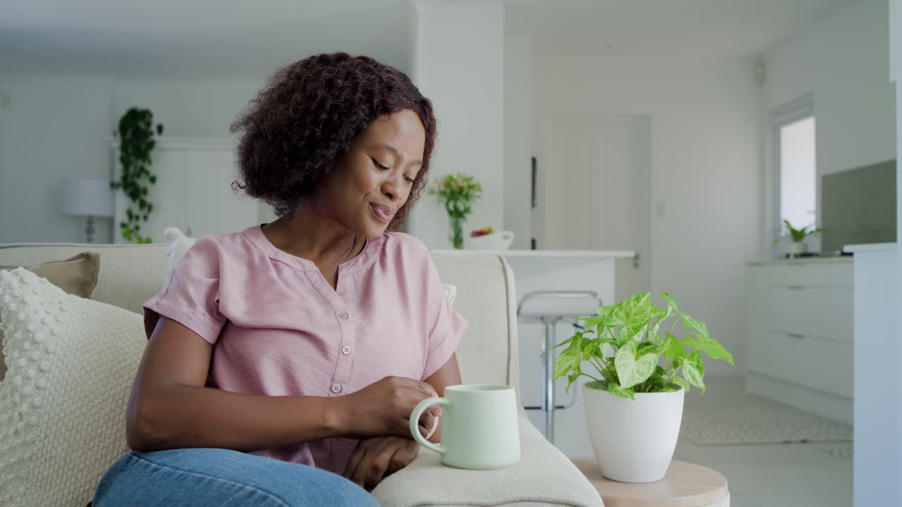 Donna africana adulta che stringe una tazza verde chiaro, sorseggiando, rilassandosi sul divano con camicetta rosa e jeans blu