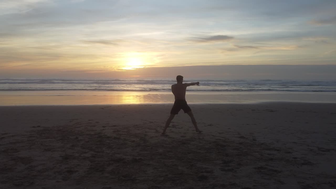 Low drone shot panning right, of a man shadow boxing on a beach in Portugal, Costa da Caparica, with an amazing background sunset.