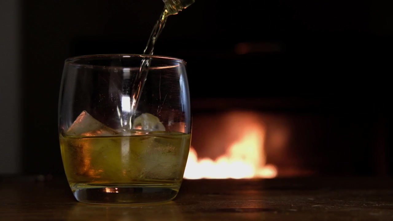 Primo piano di ghiaccio e whisky dorato versati in un bicchiere davanti a un caminetto.