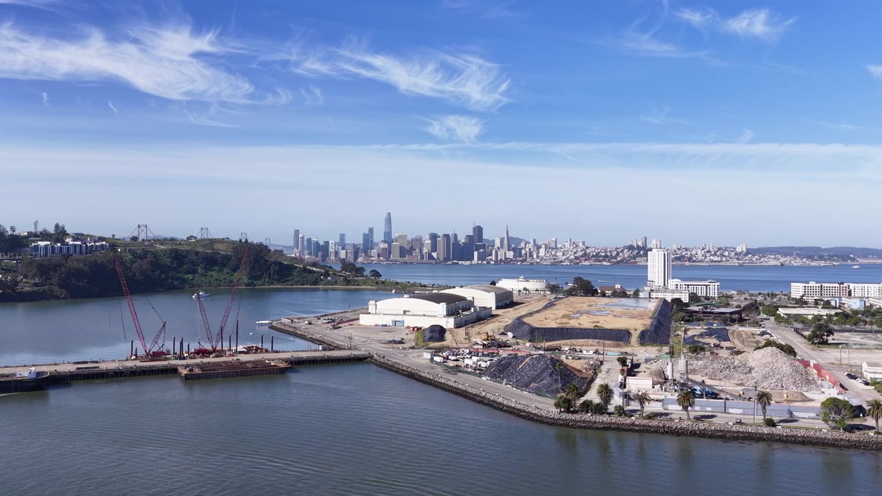 Vista mattutina con drone che sorvola Treasure Island nella baia di San Francisco, mostrando progetti di costruzione in corso con l'iconico skyline del centro città e l'isola di Yerba Buena sullo sfondo