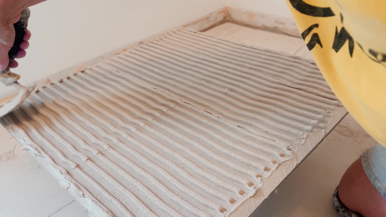 Nærbillede af en fliselægger, der spreder klæbemiddel med en tandspartel og skaber jævne riller til keramiske gulve under en indendørs renovering og professionel installation.