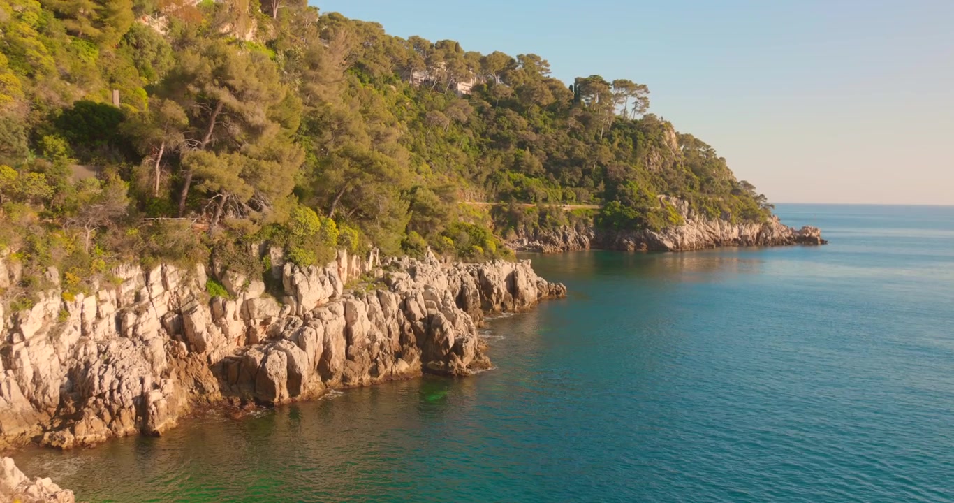 Ripresa aerea bassa che segue scogliere rocciose aspre coperte da foresta mediterranea che incontrano un mare turchese calmo.