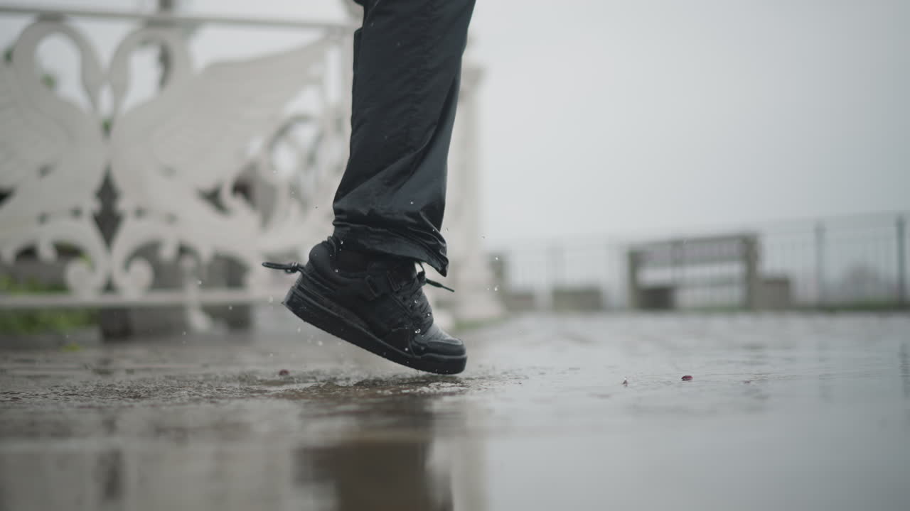 Woman splashing in puddle rainy promenade, black boots kicking water against ornate white railing under overcast sky wet pavement reflects motion and droplets, candid urban mood mixing playful energy