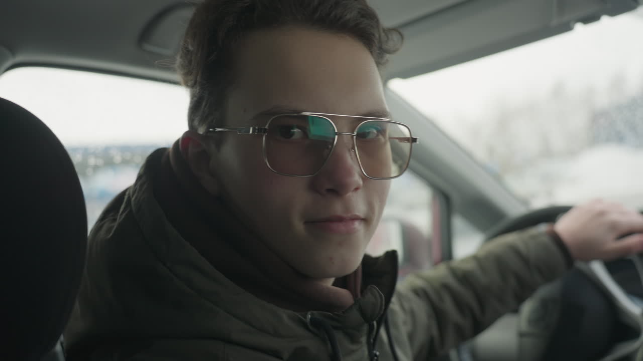close up of boy in winter jacket and sunglasses holding steering wheel turning toward camera with soft smile on face as snow falls outside car window in cold urban background