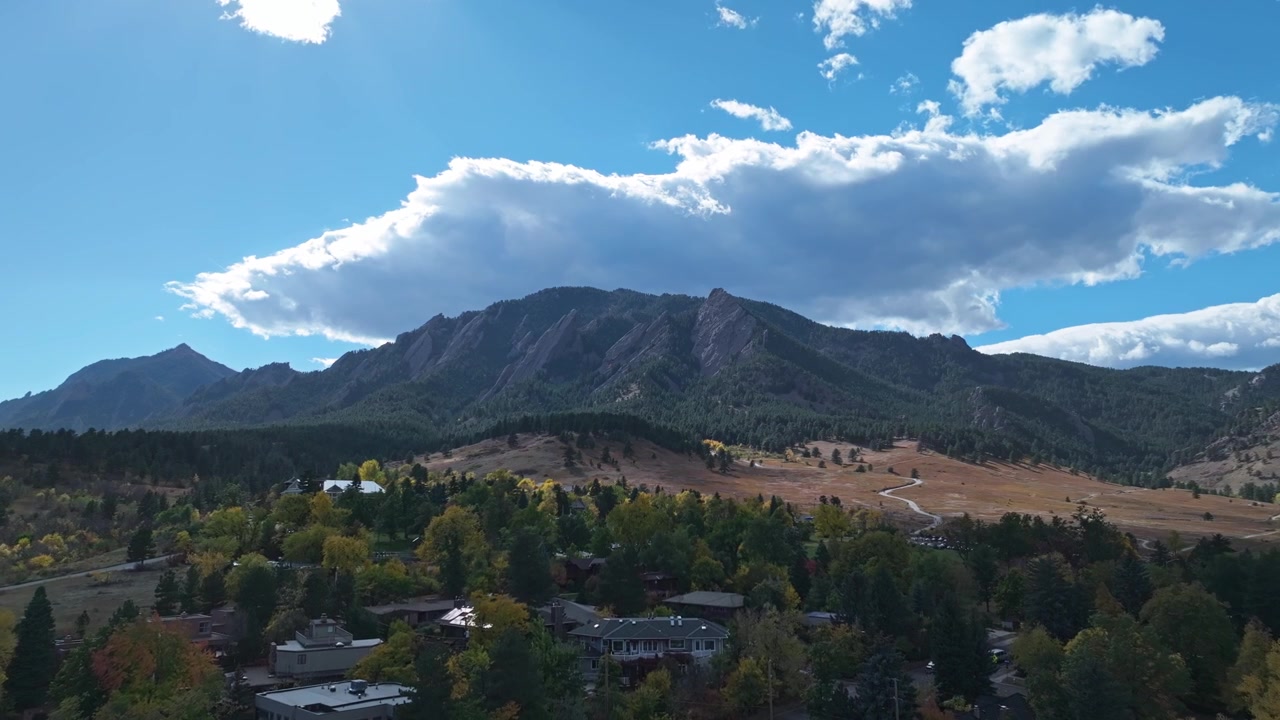 Panoramisk luftfoto af Boulder, Colorado med farverigt efterårsløv, forstadshuse og Flatirons bjergkæden i det fjerne, skygget af skyer.