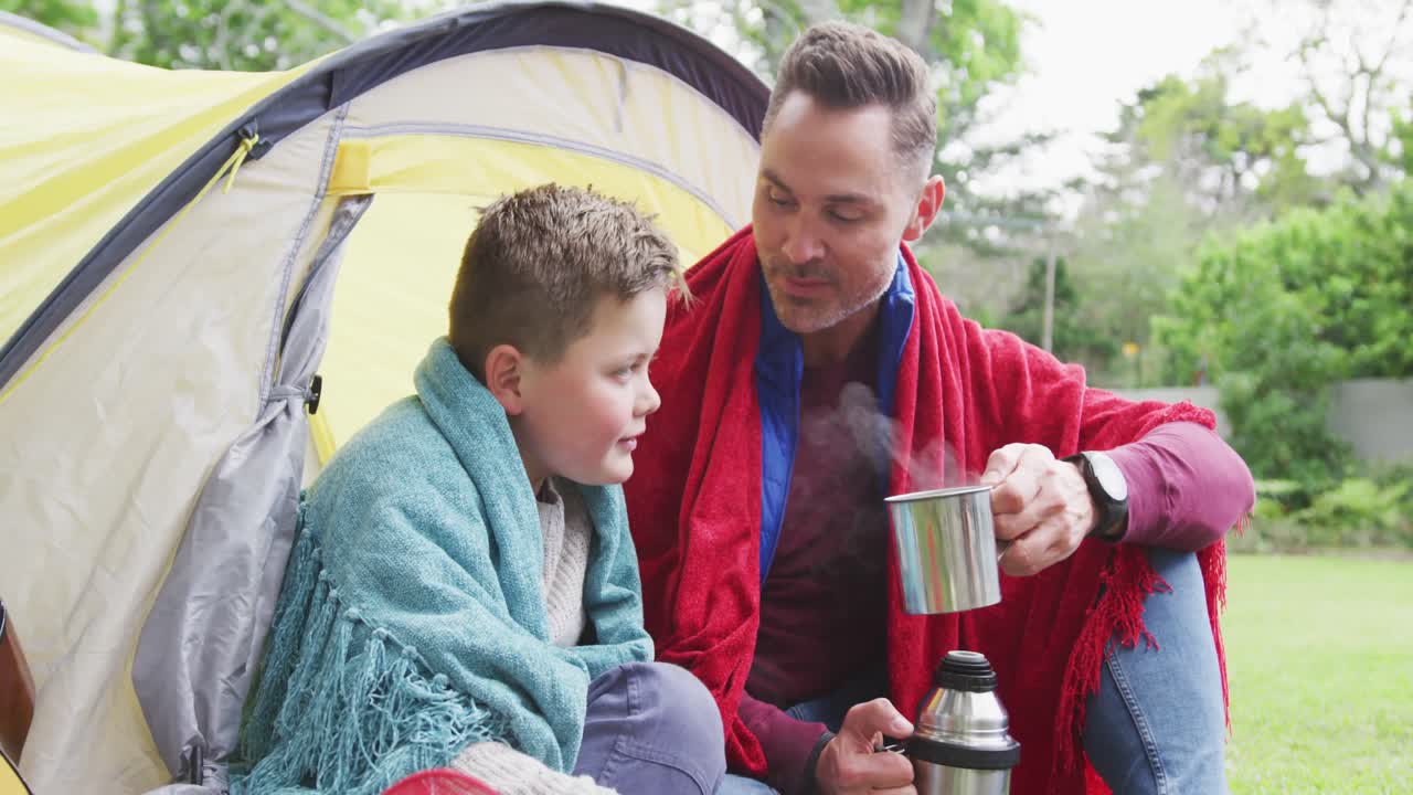 feliz padre caucásico con su hijo sentado en la tienda y bebiendo té en el jardín
