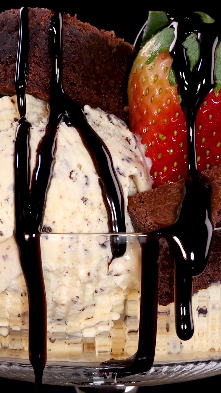 Salsa al cioccolato versata lentamente su gelato alla vaniglia con gocce di cioccolato, pezzi di brownie e fragole fresche in una ciotola di vetro, sotto un'illuminazione da studio drammatica.
