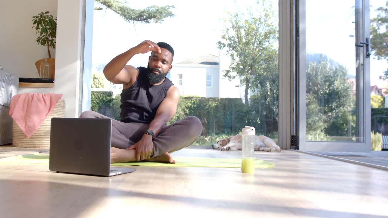 hombre afroamericano enfocado practicando yoga usando una computadora portátil en casa soleada, cámara lenta