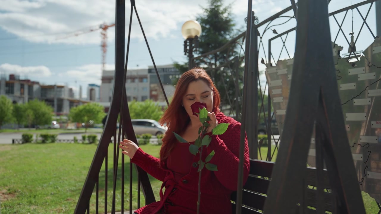 Donna con maglione rosso siede su un'altalena all'aperto, tenendo e annusando una singola rosa. La scena è ambientata con una luce diurna soffusa, con erba verde, alberi, edifici distanti e auto di passaggio visibili.
