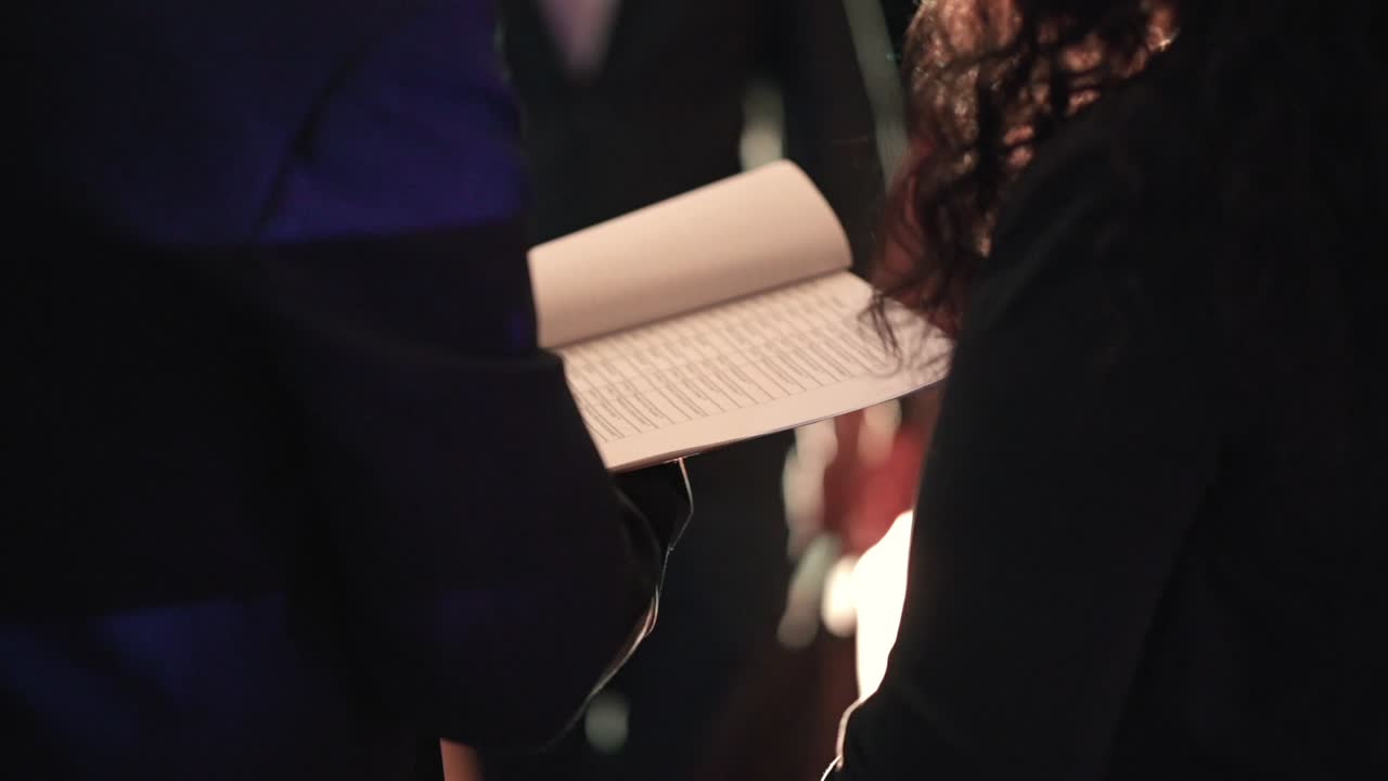 Persona che tiene in mano una lista invitati stampata mentre verifica i nomi all'ingresso dell'evento con sfondo a bassa luminosità e profondità di campo ridotta durante il controllo accessi di una reception organizzata, primo piano.