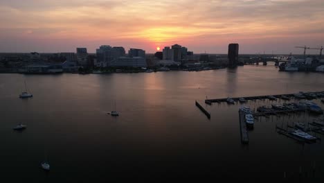 flying a drone along a marina pier with the boats in the foreground and the norfolk virginia waterfront in the background with the rising sun peaking out from behind the buildings as it moves