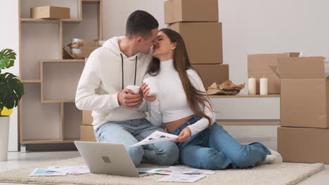 Young-Couple-In-A-New-House-Sitting-On-The-Carpet-With-Laptop-Choosing-Colours-For-Decoration-While-Drinking-Coffee