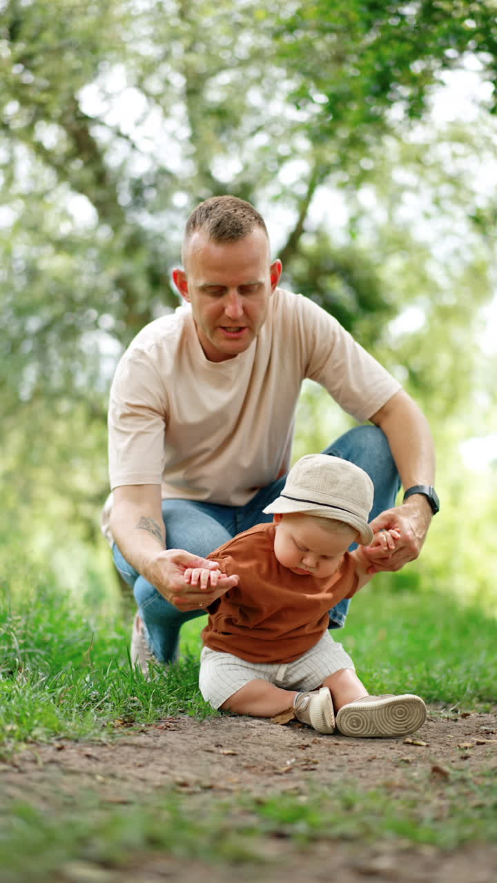 Dad sits squatted behind his son sitting on the ground. Baby boy looks at the ground trying to take a leaf. Vertical video.