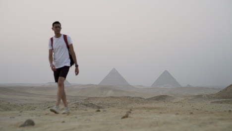 young man walks towards camera in desert with giza pyramids in background