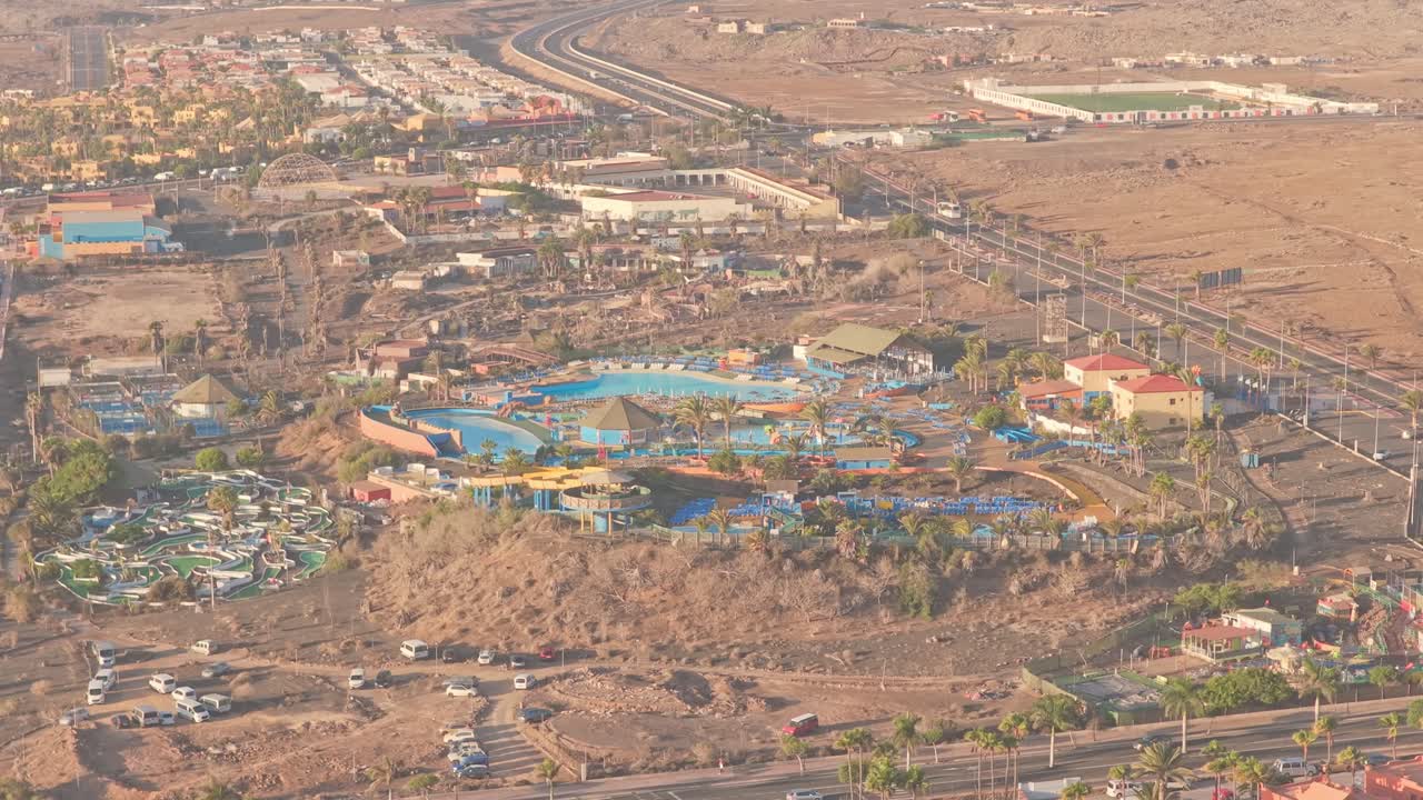 Aerial view revealing Acua Water Park's colorful swimming pools and slides nestled among hotels and arid terrain of Fuerteventura, Canary Islands, under bright sunlight