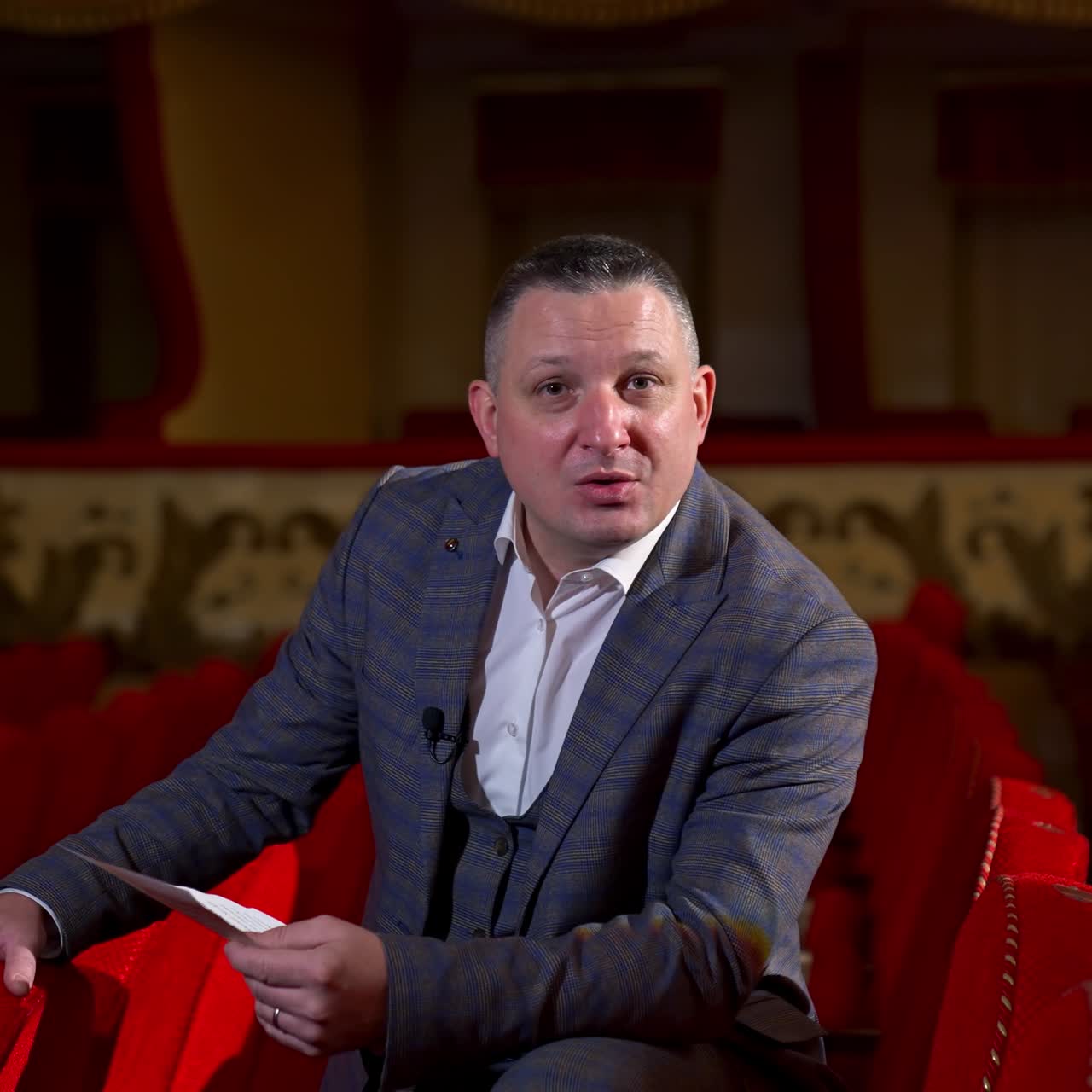 Host announces future theater season. Actor is sitting in empty theater on red velvet chair. Classic theater interior in red colors