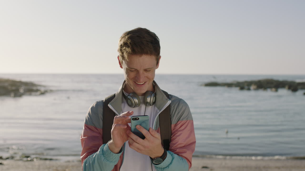 retrato de un joven enviando mensajes de texto navegando usando el teléfono de pie en la playa sonriendo feliz