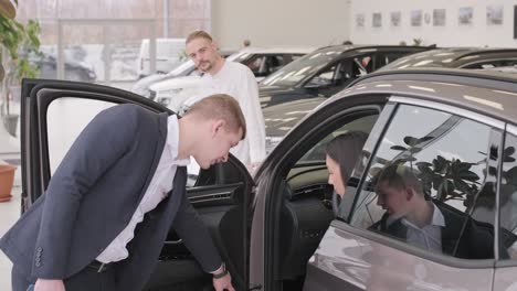 young car salesman showing to young couple new automobile at dealership salon.