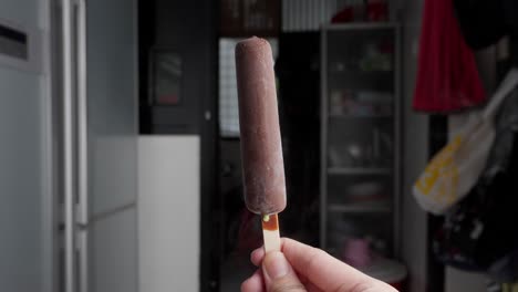 Hand-holding-chocolate-ice-cream-popsicle-against-blurry-urban-background,-focus-on-foreground