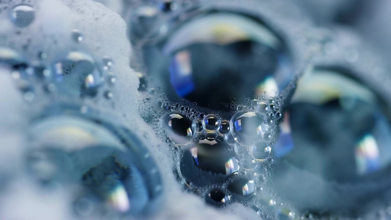 Denso grappolo di bolle di sapone e schiuma spumosa in primissimo piano, con piccole forme sferiche, superfici lucide e riflettenti, sottili ponti liquidi, luce blu soffusa e una fine texture astratta.