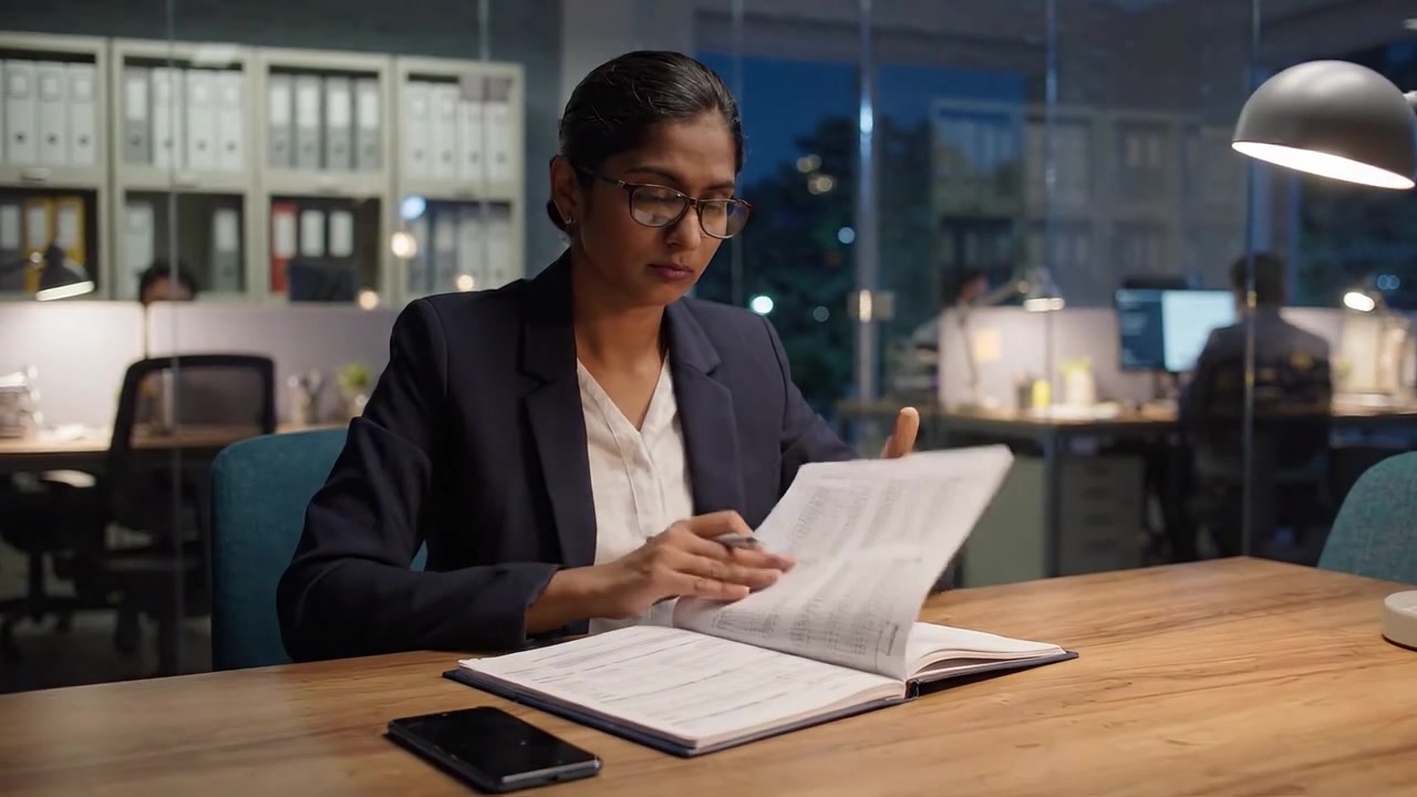 Focused Professional in Smart Attire Concentrates on Important Work at a Well-Lit Office Table During Late Evening Hours, Exhibiting Dedication and Precision
