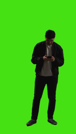 Vertical-Video-Full-Length-Shot-Of-Young-Man-Text-Messaging-On-Mobile-Phone-Standing-Against-Green-Screen-2