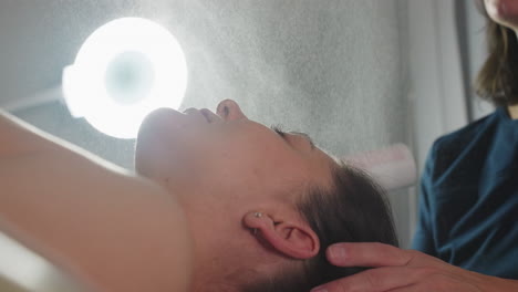 therapist hand holds spray bottle misting nourishing serum onto face of spa guest lying under bright lamp while supporting head for refreshing hydration and skin soothing during facial treatment