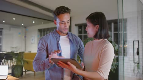 focused diverse male and female colleague standing talking and using tablet at night in office