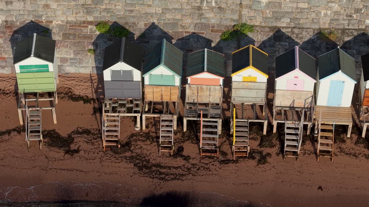 Aerial drone pans across bright Torquay beach huts. View highlights colorful doors and rooftops above sandy beach. Perfect seaside vacation stock clip with holiday, leisure, and relaxation themes
