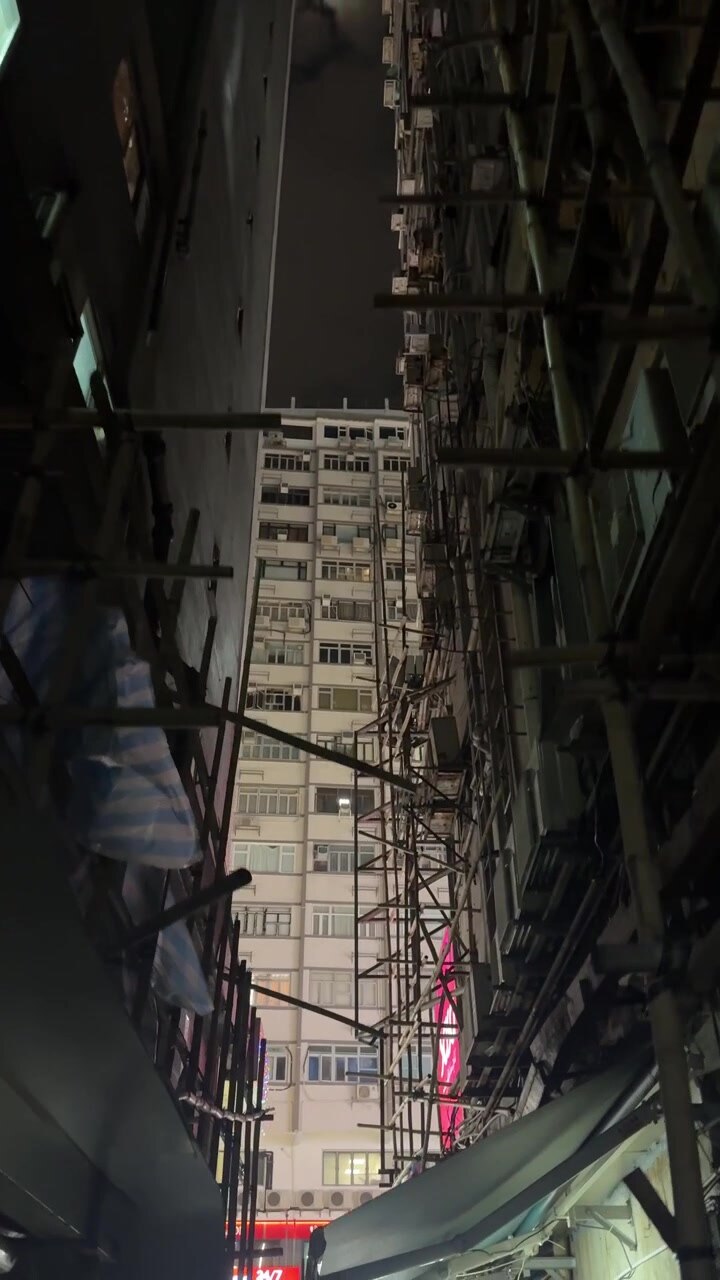 Vista nocturna vertical mirando hacia un callejón estrecho enmarcado por andamios de bambú y edificios de gran altura en Hong Kong.