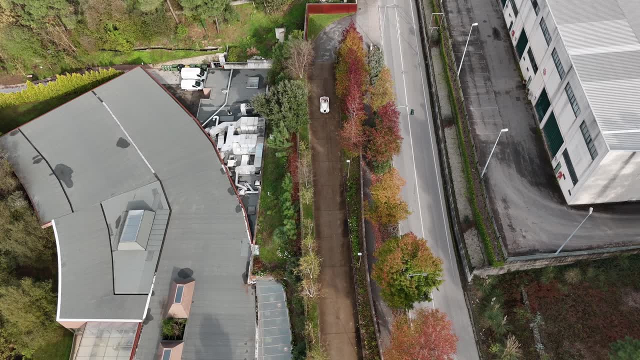 Aerial - classic white car on tree-lined road between industrial buildings