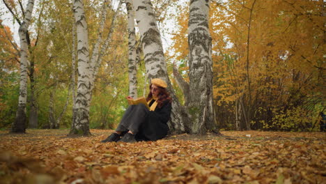 tourist enjoying book in solitude wears yellow beret scarf around neck, deeply focused on book in beautiful autumn park, surrounded by golden leaves and birch trees