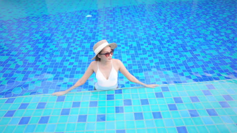 Frau-In-Weißer-Monokini-Sonnenbrille-Und-Sonnenhut-Im-Swimmingpool-Mit-Gefliestem-Boden,-Der-Sich-An-Die-Grenze-Anlehnt,-Zeitlupe,-Erhöhte-Ansicht