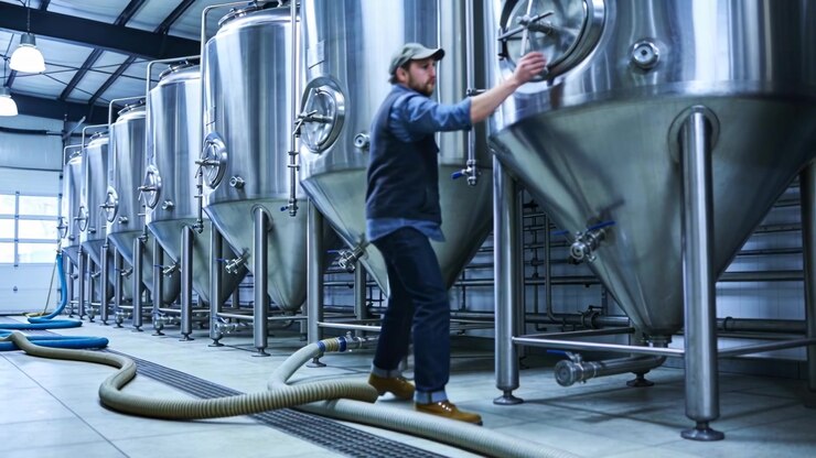 Connecting brewer moving hose, turning handwheel by conical tanks, in cap and vest for transfer
