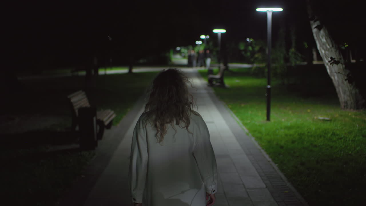 Back view of girl wearing white coat walking alone at night in quiet park walkway, soft lamp glow illuminating path, distant figures approaching in background