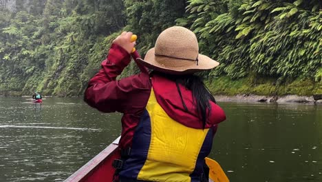 A-woman-with-black-hair-wearing-a-sun-hat,-red-jacket-and-yellow-life-vest-canoes-down-the-lush-Whanganui-River-on-a-rainy-day