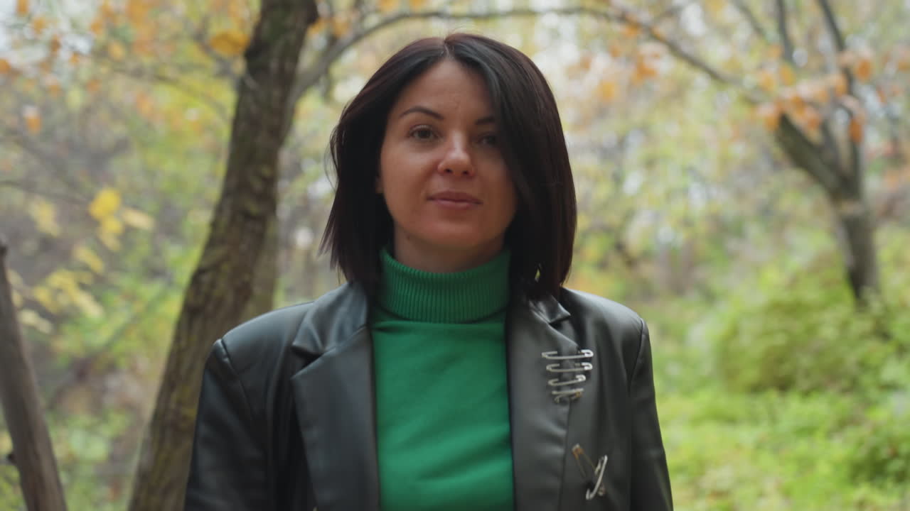 Mujer segura de sí misma con chaqueta de cuero caminando por un bosque otoñal, con un jersey de cuello alto verde, pantalones de cuero y un abrigo largo en un sendero frondoso; paseo tranquilo y, a continuación, pausa con los brazos levantados; primerísimos planos que muestran suavidad