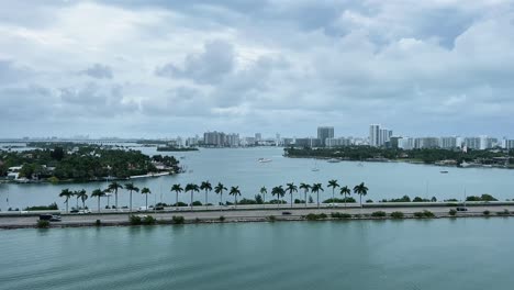 Tilt-up-shot-of-a-busy-highway-surrounded-by-water-in-Miami-Florida-with-tropical-palm-trees,-boats,-and-sky-scrapers-in-the-distance-on-an-overcast-day