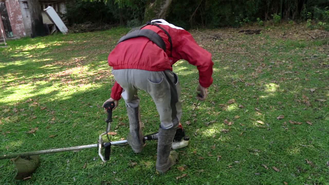 A gardener in a red jacket pulling the starter cord of a string trimmer on a green lawn. Slow motion