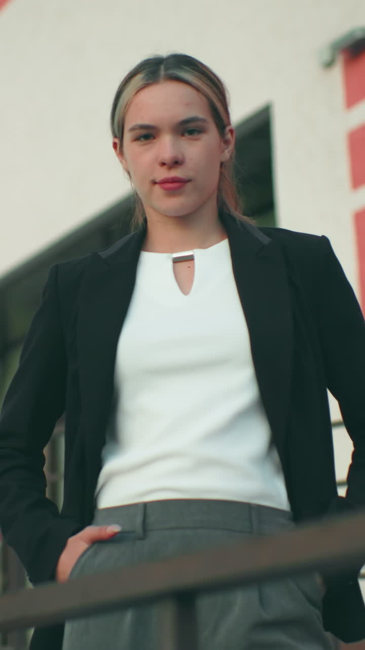 Confident lady posing with warm smile beside iron railing in front of office building featuring striped walls under clear sky, hands in pockets, exuding elegance and professionalism in urban setting