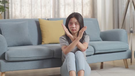 young asian woman with bruise on body looking into camera showing hands cross gesture to stop violence while sitting on the floor at home