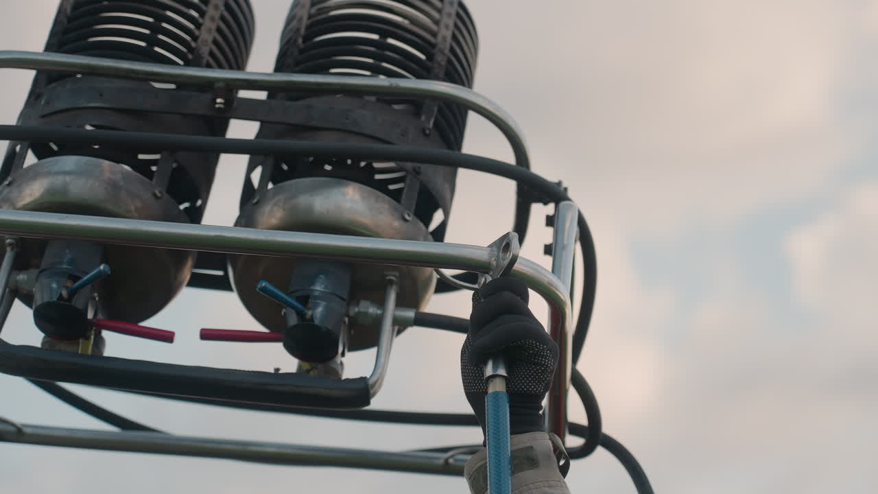 gloved hands adjusting metal burner assembly onto support frame of hot air balloon under pale sky preparing for flight in open field setting with focus on manual installation