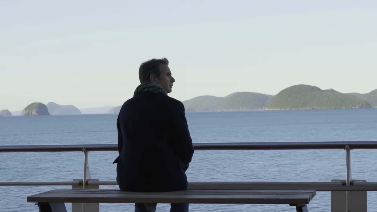 White Man Sitting Bench By Sea, Distant Island Horizon And Calm Water, Cool Morning Light, Navy Coat