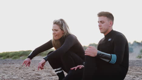 couple wearing wetsuits sitting on surfboard before getting up and walking along beach