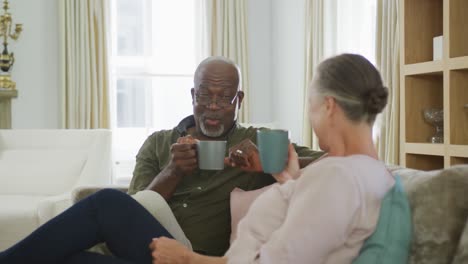 Glückliches-älteres,-Vielfältiges-Paar,-Das-Hemden-Trägt-Und-Kaffee-Im-Wohnzimmer-Trinkt