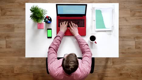 young man in glasses typing on laptop and using phone with chroma key, topshot, sitting behind desk