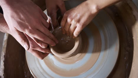 Close-Up-Of-Male-Teacher-Helping-Woman-Sitting-At-Wheel-In-Pottery-Class